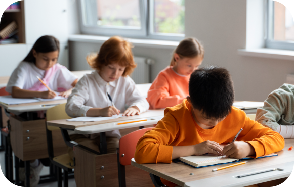 Young students diligently write at individual desks in a brightly lit classroom.