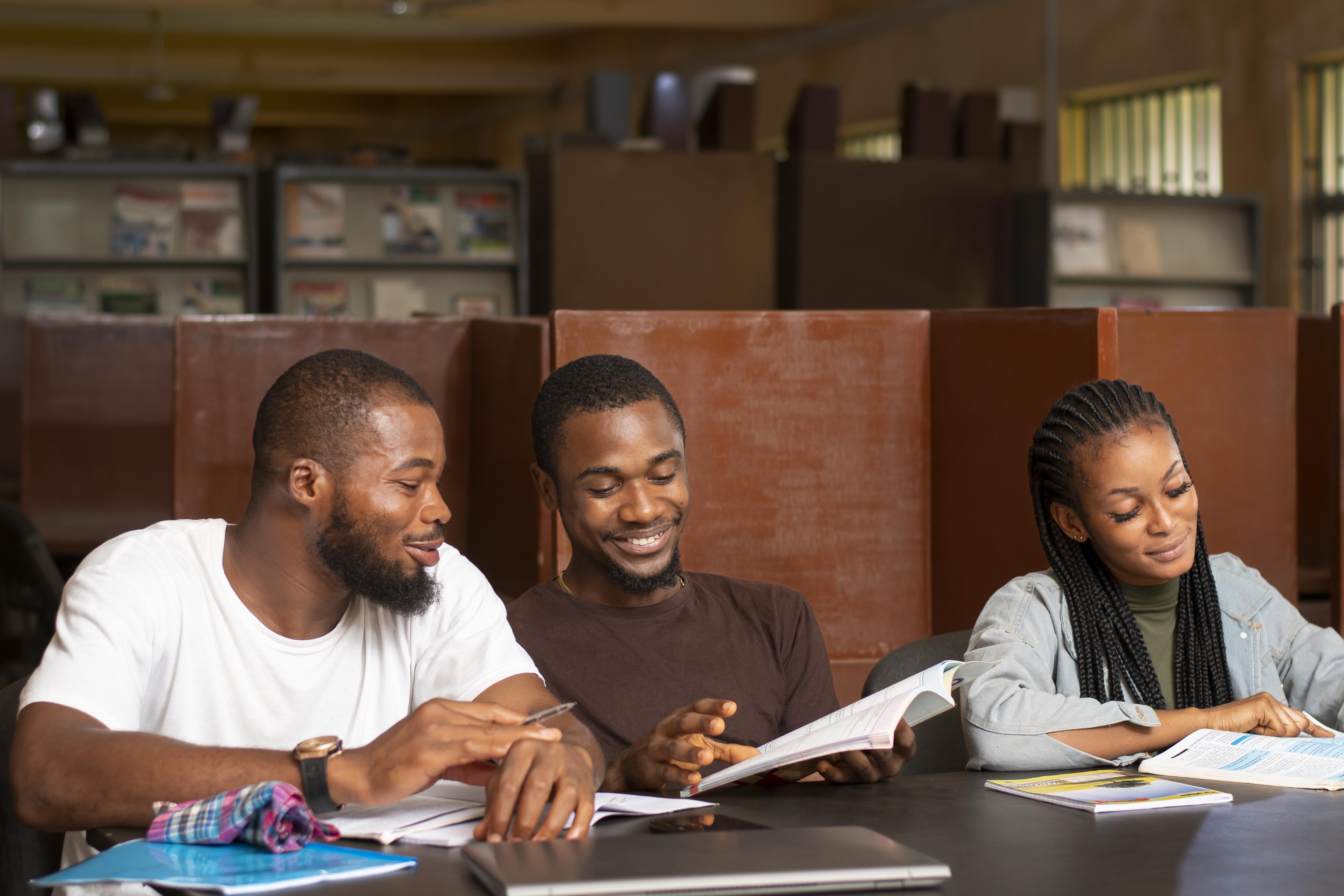 Three students studying together at a desk with books and a laptop in a library.