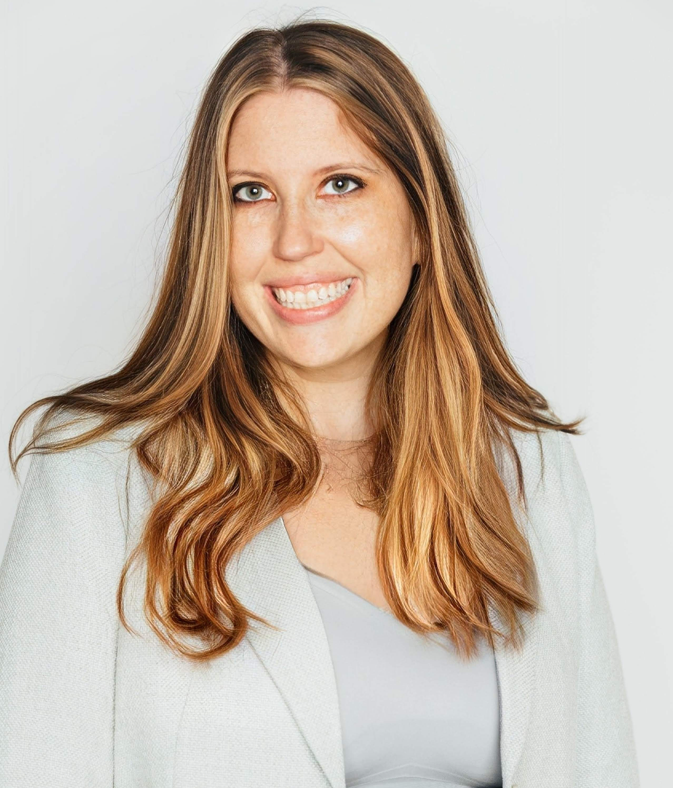  A portrait of a smiling woman with long, wavy blonde and brown hair, wearing a light gray or pale blue blazer over a solid top, against a white background.