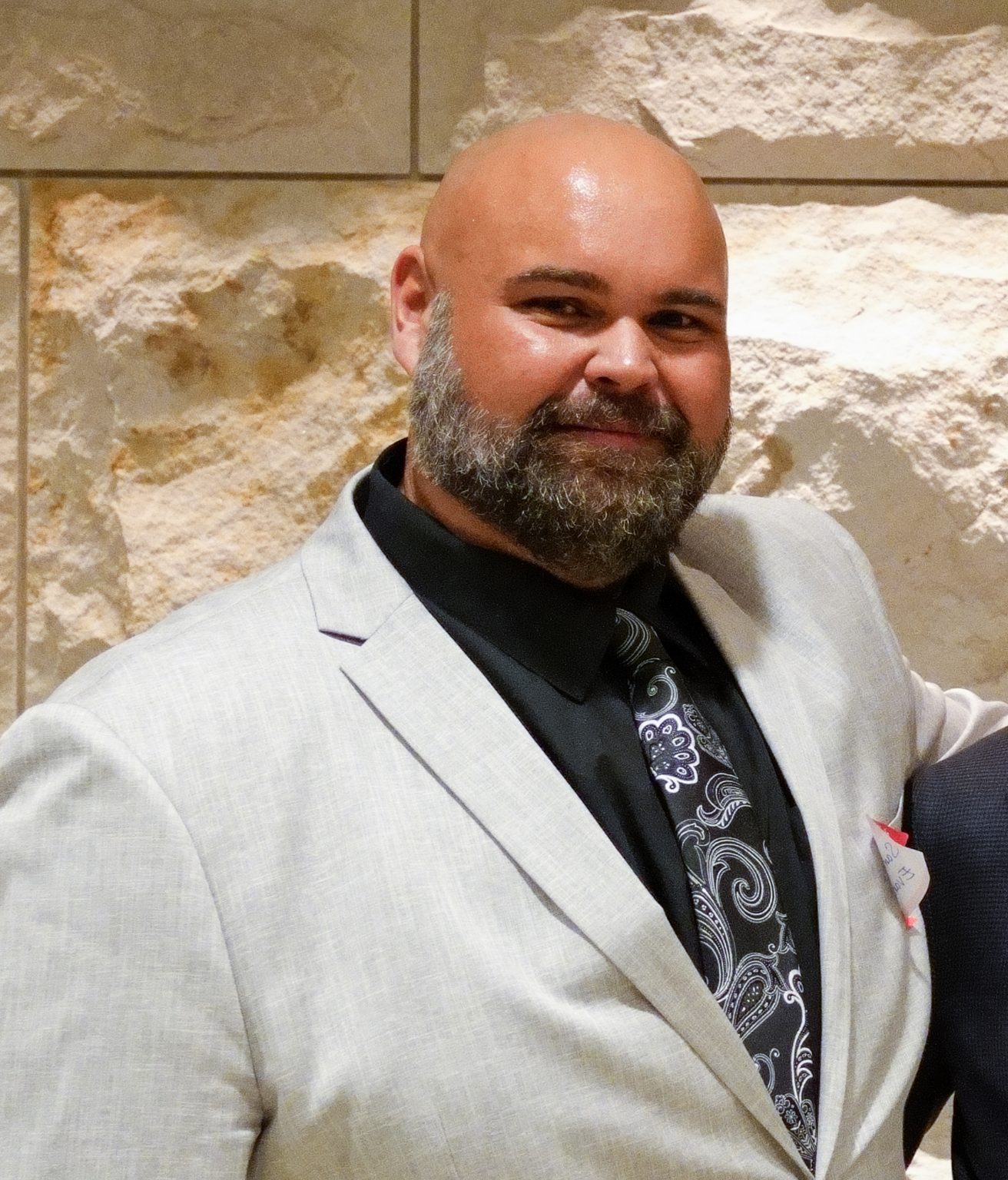  A man with a bald head and a full beard smiles, wearing a light gray suit jacket, a black shirt, and a black and white paisley tie, standing against a textured light stone wall.