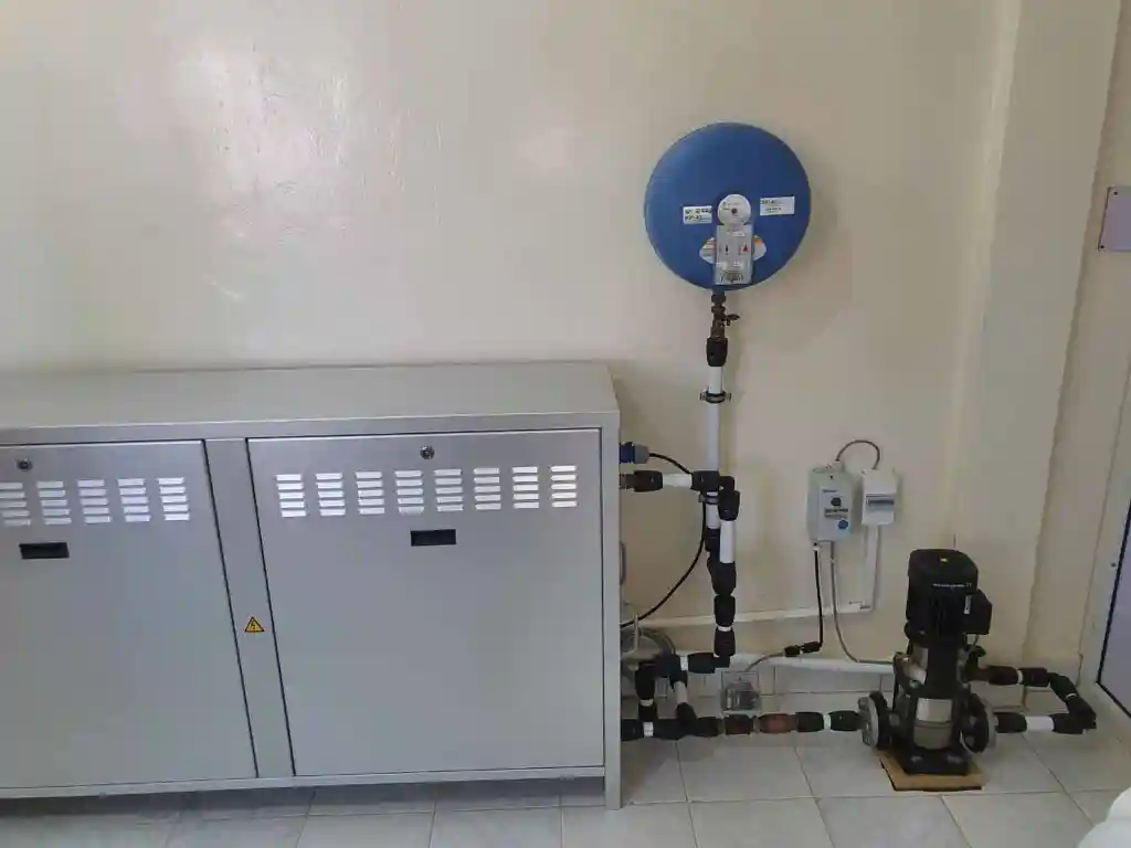 Metal cabinet with ventilation panels next to a blue circular water meter and connected pipes on a tiled floor against a beige wall.