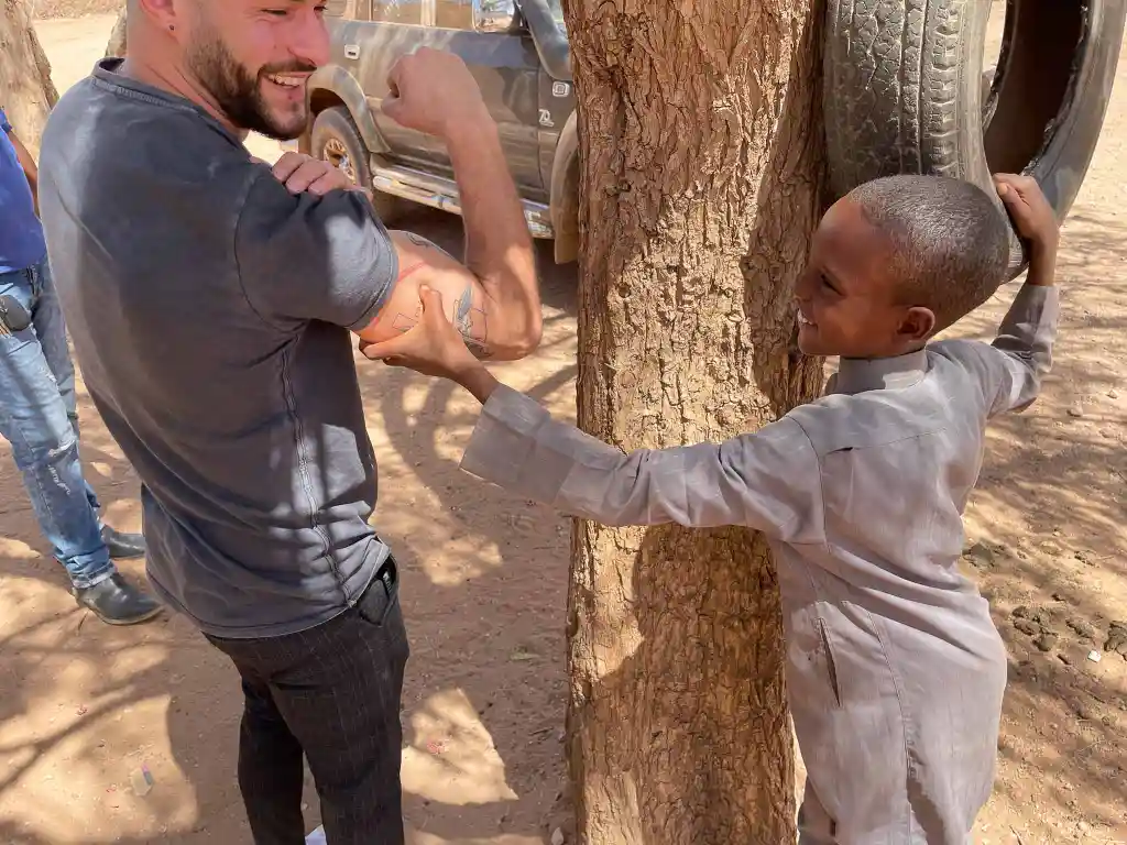 A smiling boy holding a tire swing is playfully pressing the arm of a man who is flexing his bicep outdoors.