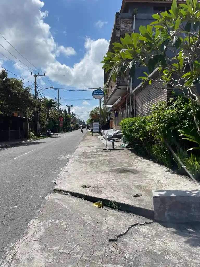 Sunny daytime street view with a cracked sidewalk, a brick building on the right, and greenery along the path.