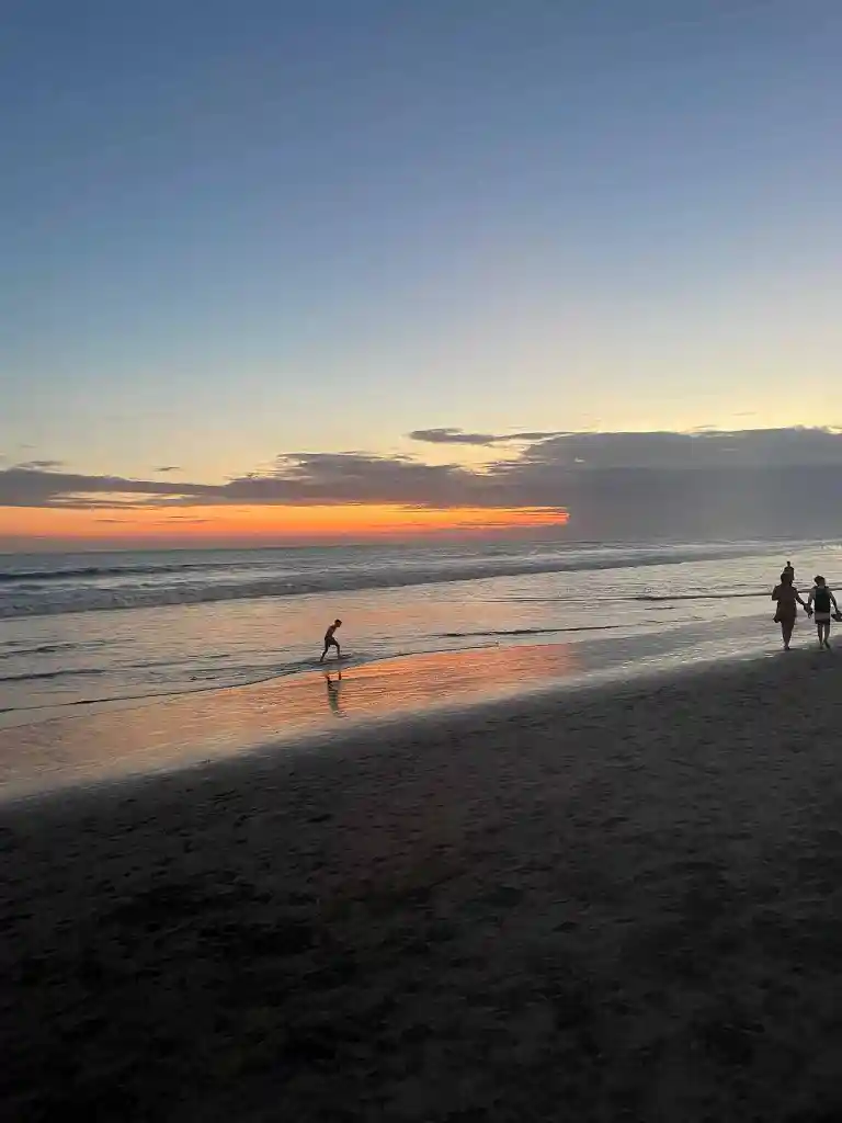 Silhouettes of a child playing near the ocean's edge and two people walking on a beach at sunset with an orange and blue sky.