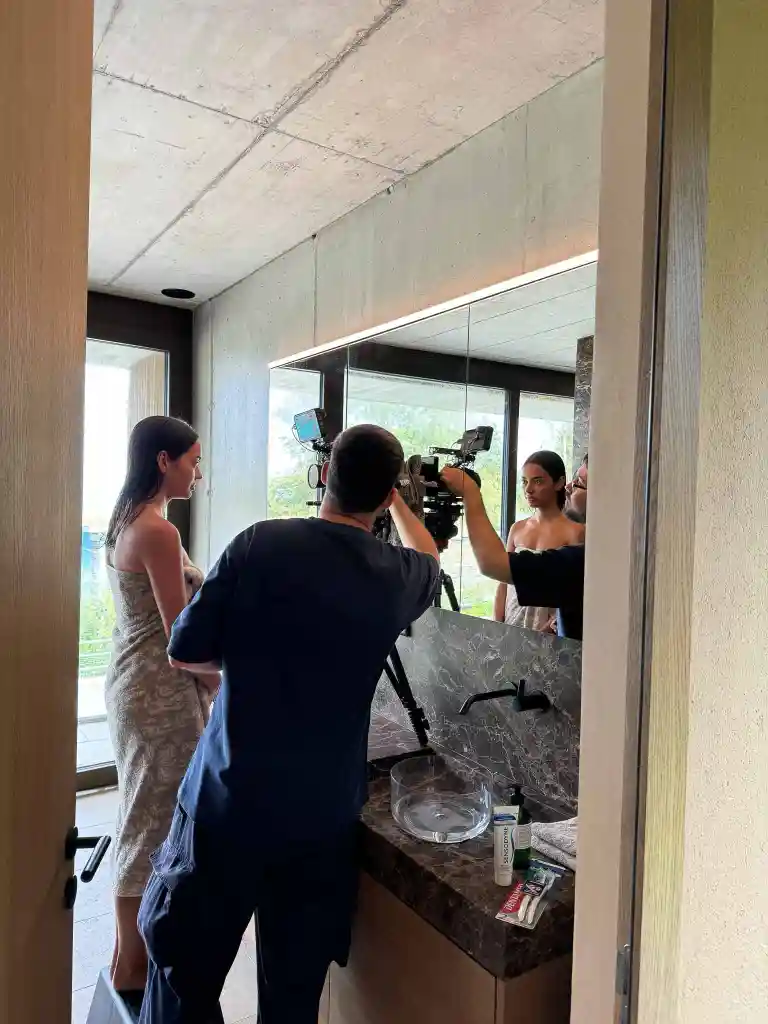 Photographer adjusts camera on tripod in front of a woman wrapped in a towel standing by a bathroom sink with a large mirror.