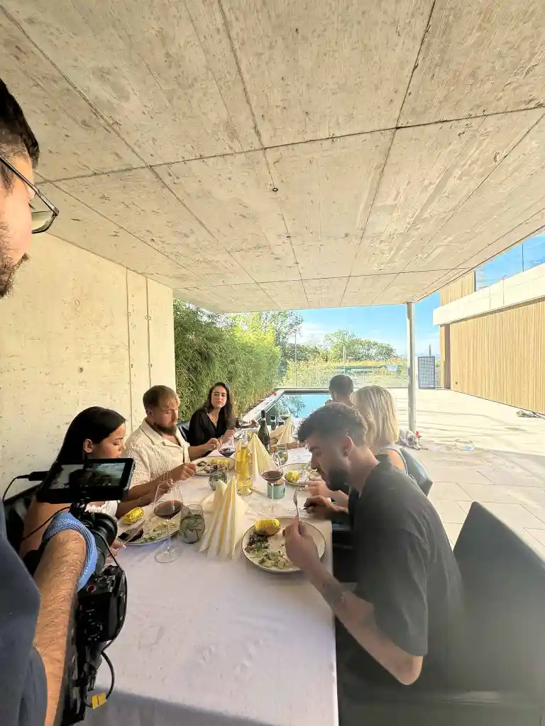 Group of seven people dining outdoors under a concrete canopy with wine and food on the table.