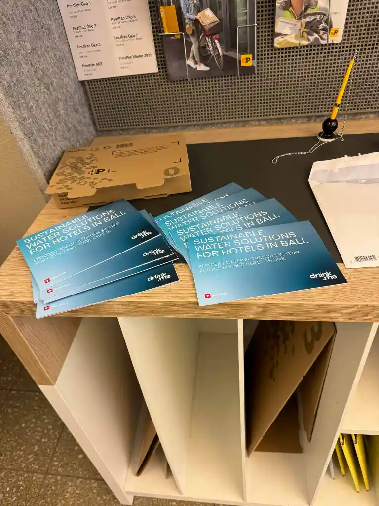 Brochures titled 'Sustainable Water Solutions for Hotels in Bali' arranged on a wooden counter with other packaging materials and a pen attached by a chain.