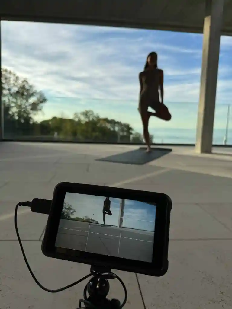 Camera on tripod recording a person doing a yoga tree pose near a large window with an outdoor view.
