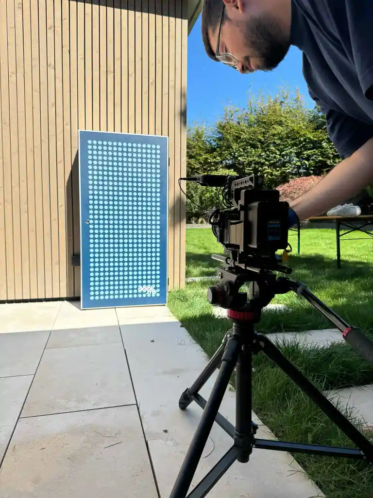 A man adjusting a professional video camera on a tripod outside, aiming at a wooden wall with a blue rectangular panel featuring a grid of white dots and the word 'drink.'