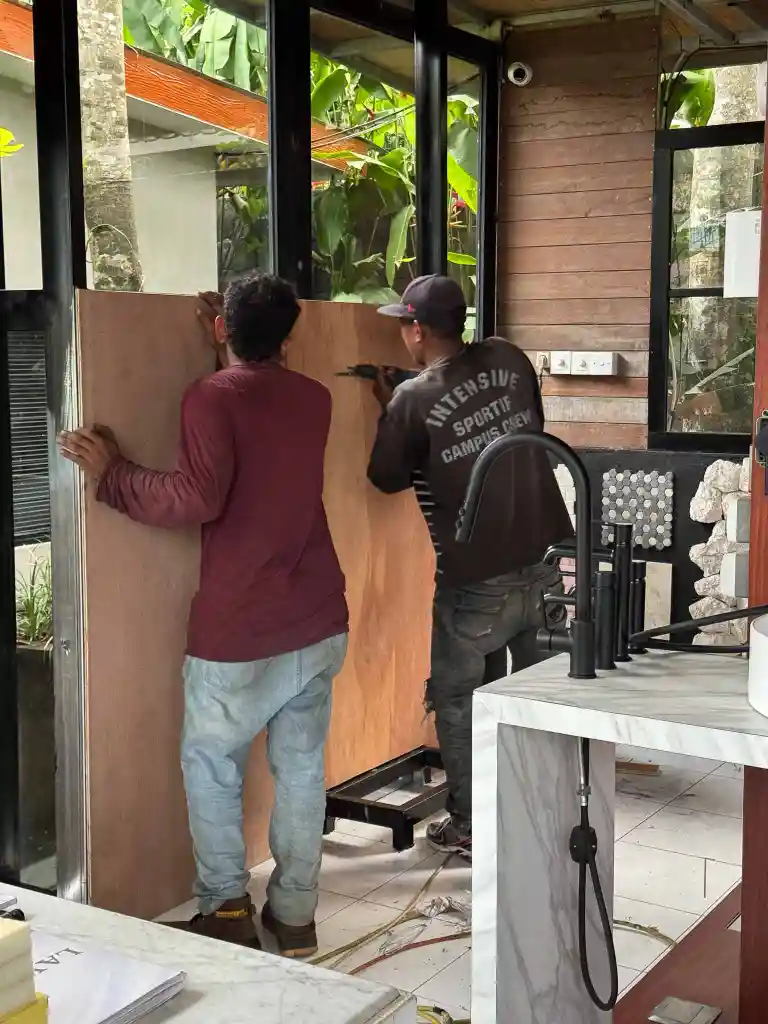 Two men installing a large wooden panel on a glass door frame inside a room with marble countertops and black faucets.