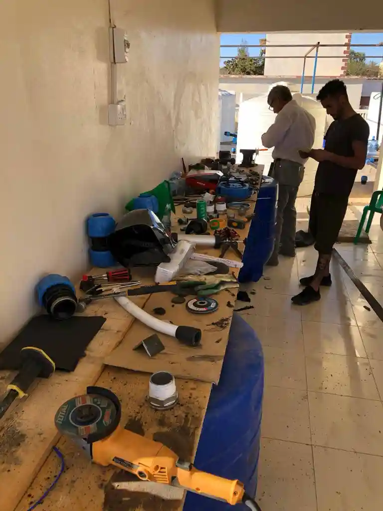 Two men standing next to a workbench filled with plumbing tools and equipment in a workshop area.