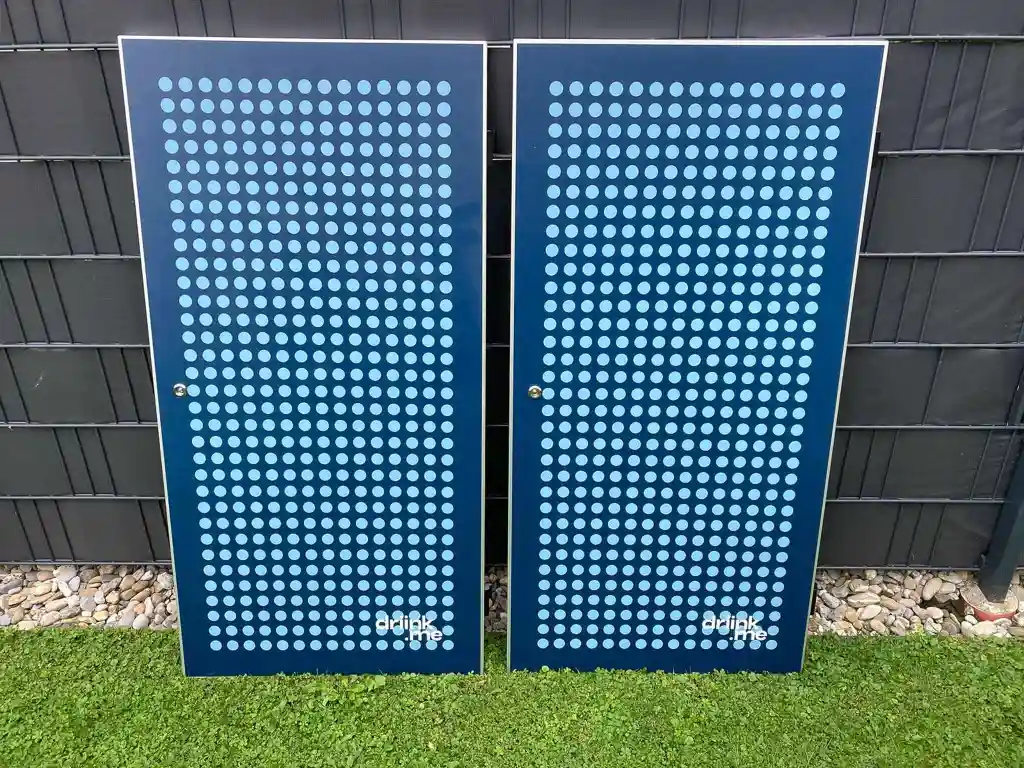 Two blue rectangular boards with a grid of light blue dots and the text 'drink me' at the bottom, leaning against a dark metal fence on grass.