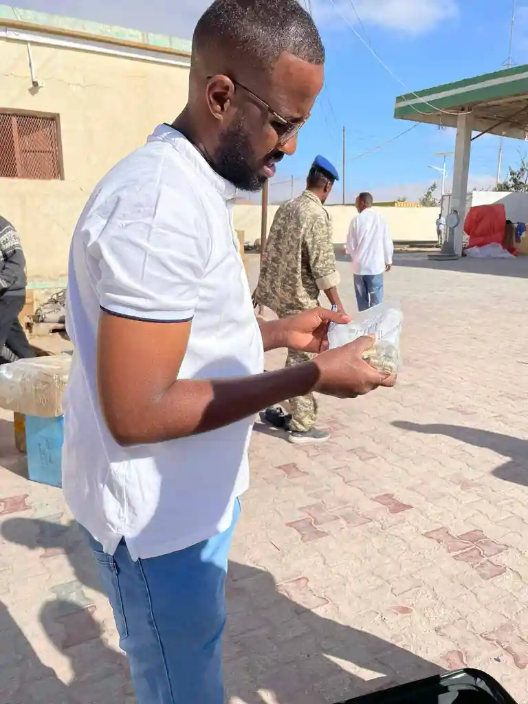 Man in white shirt and blue jeans outdoors holding a clear plastic bag with objects inside, with other people and buildings in the background.