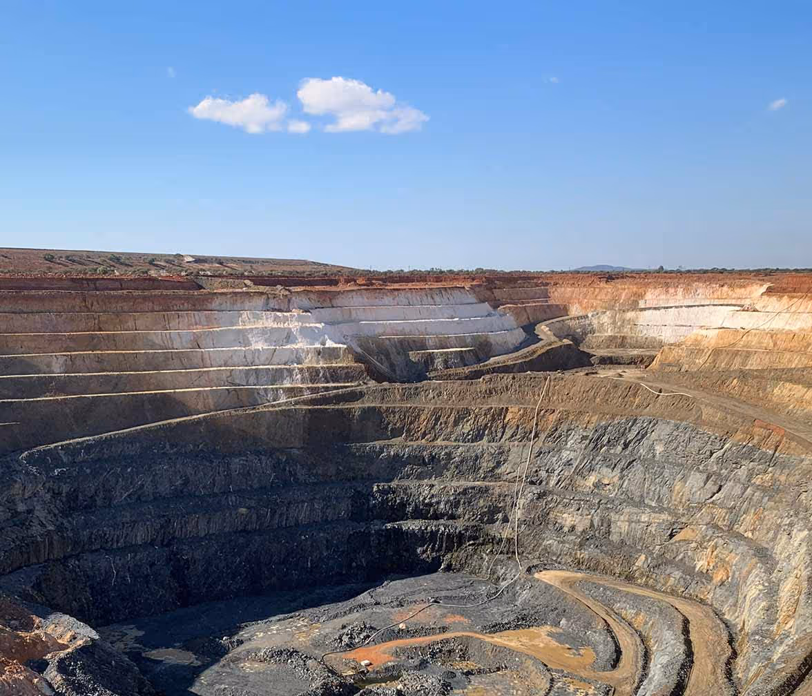 Open-pit mine with terraced layers of exposed earth under a clear blue sky with a few clouds.
