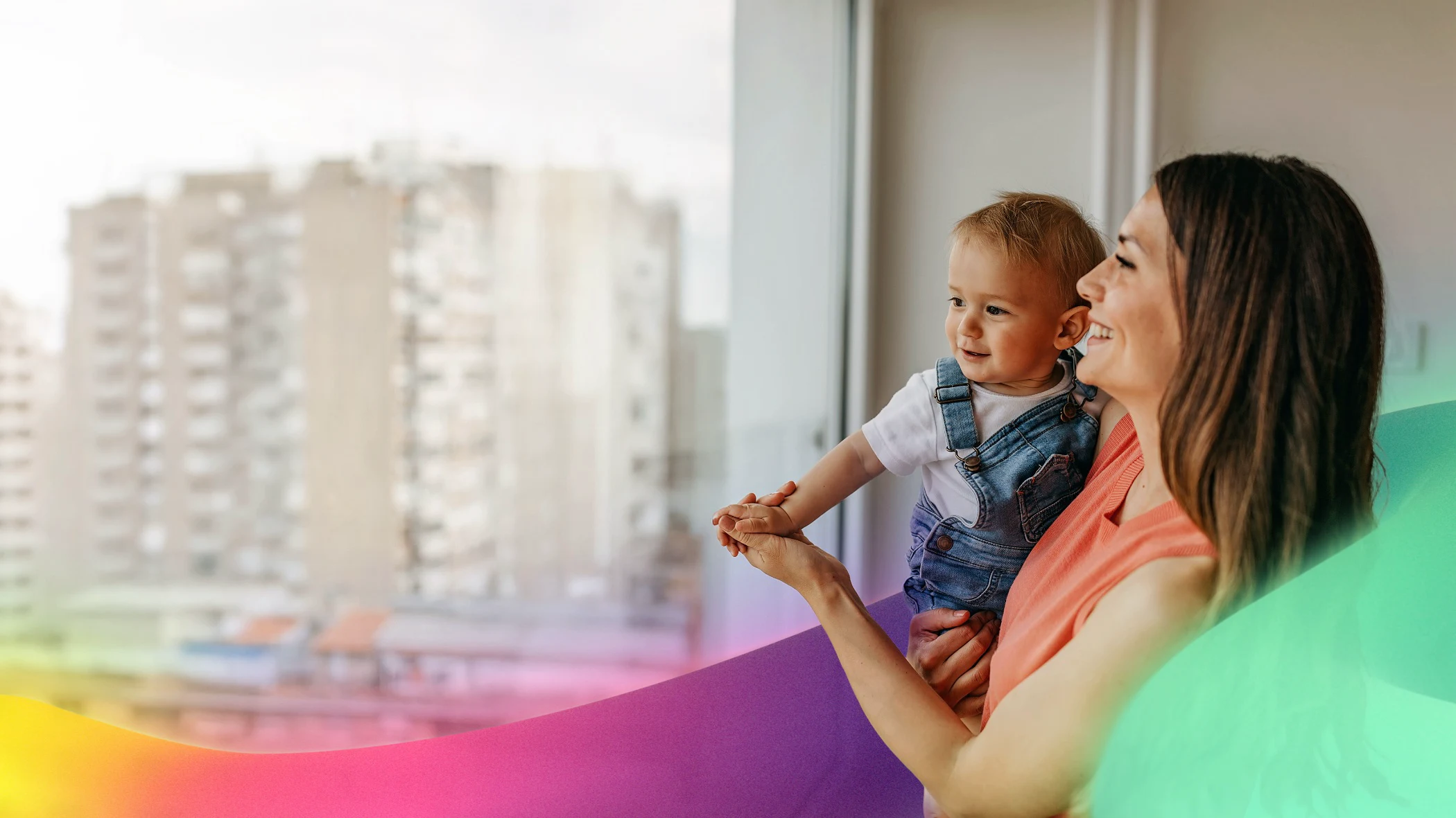 Smiling woman holding happy baby by window with city buildings outside