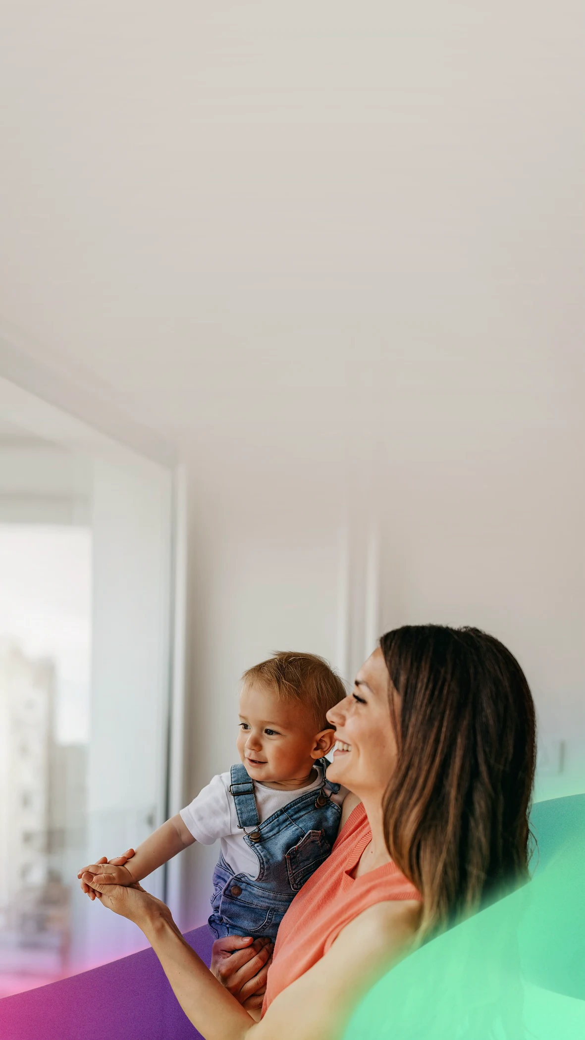 Smiling mother holding baby in denim overalls by bright window