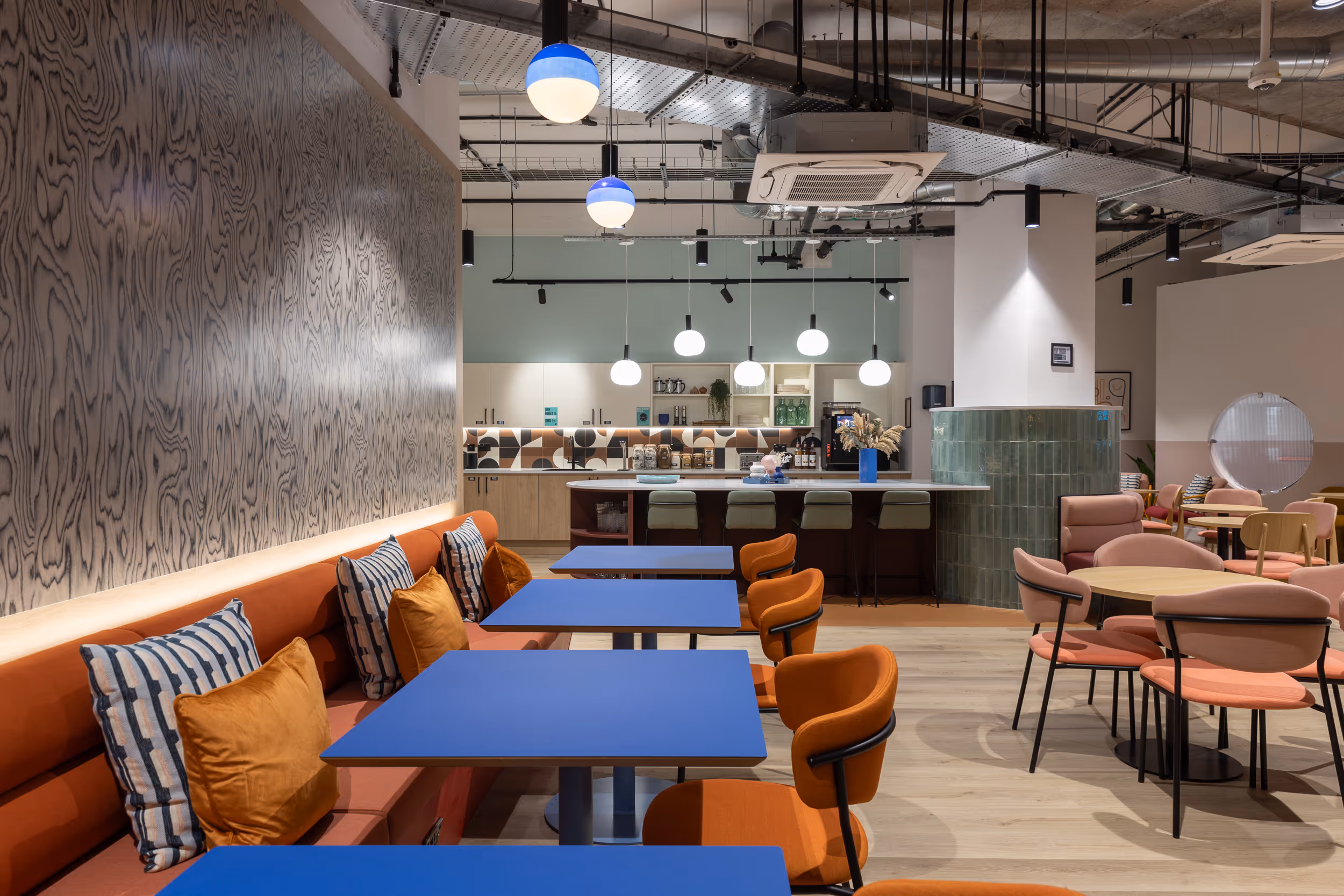 Modern office breakout space with blue tables, orange seating, and kitchen area