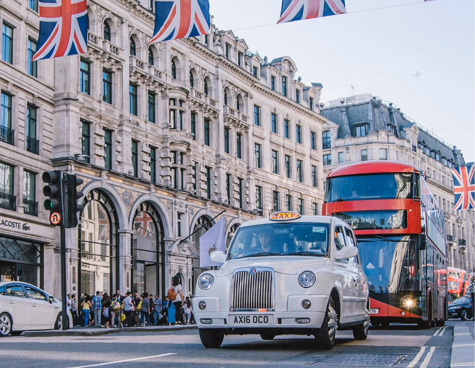 White London taxi and red double-decker bus on a street with historic buildings and Union Jack flags overhead.