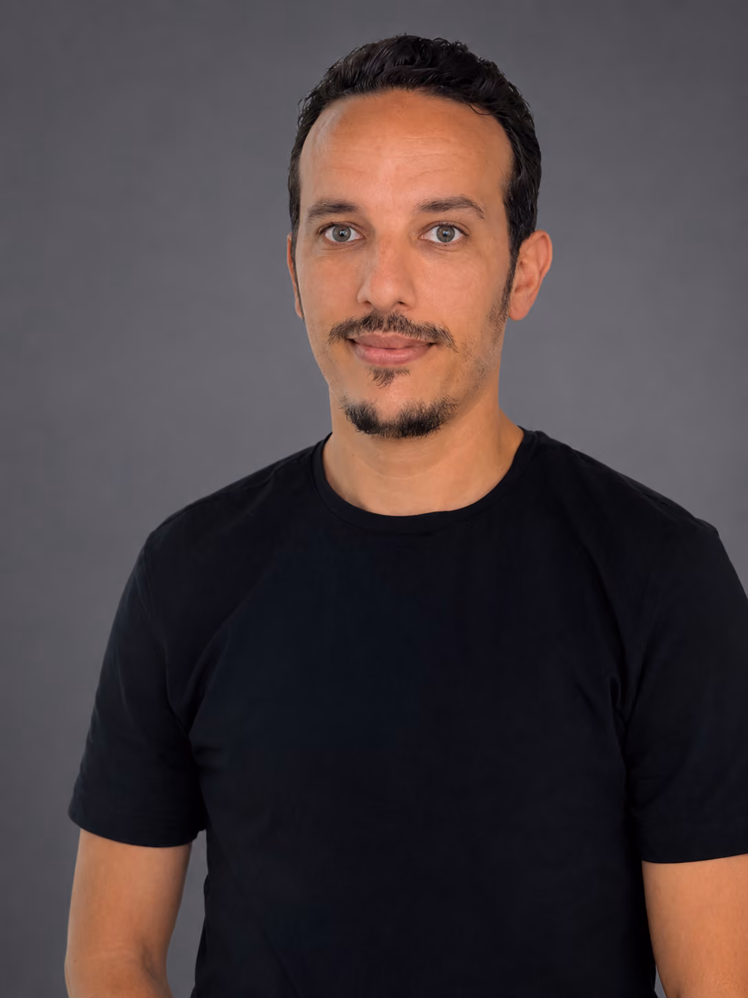 Man with short black hair and goatee wearing a black t-shirt against a gray background.