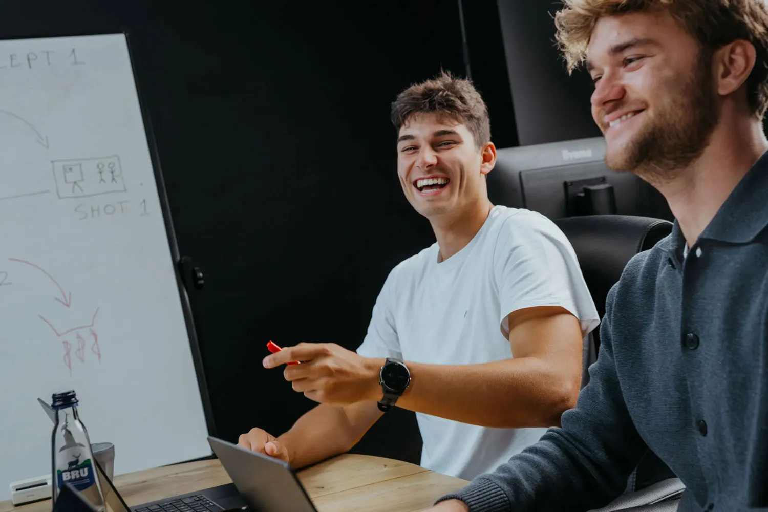 Two young men sitting at a table with laptops, smiling and laughing during a discussion near a whiteboard with sketches.