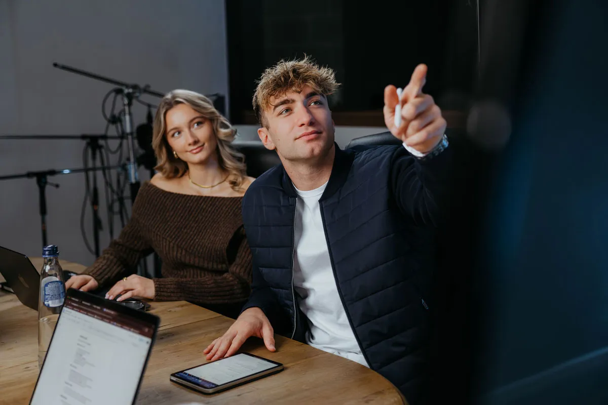 A man points at a screen while a woman watches attentively during a meeting at a wooden table with laptops and a tablet.