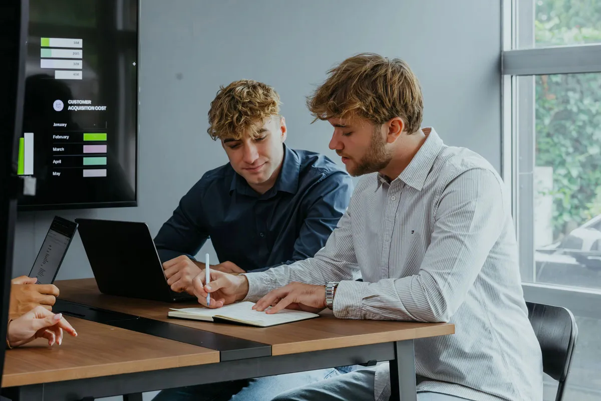 Two young men sitting at a table with laptops, one writing in a notebook, discussing work in a modern office.