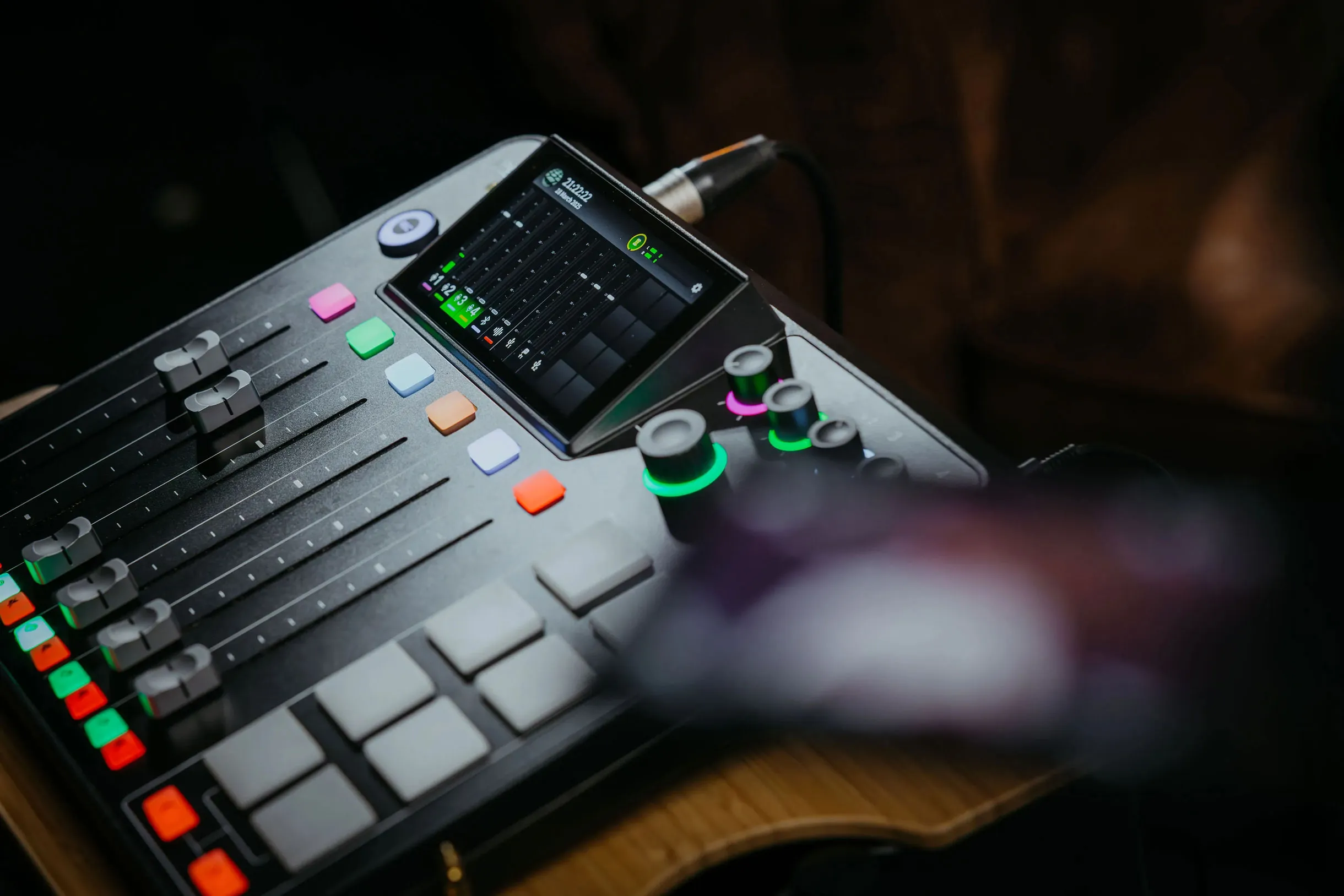 Close-up of an audio mixing console with faders, colored buttons, knobs with green lights, and a digital screen showing sound levels.