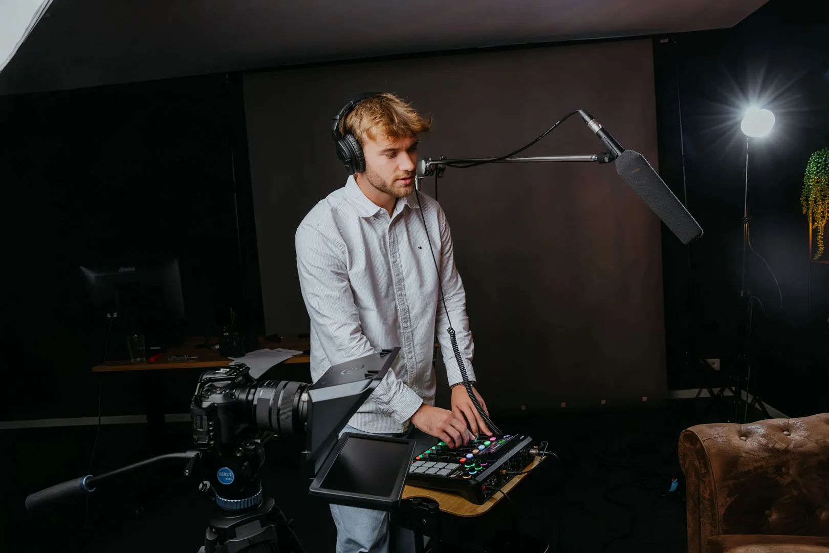 A man wearing headphones operates a sound mixer in a dimly lit studio with a microphone boom and camera setup.