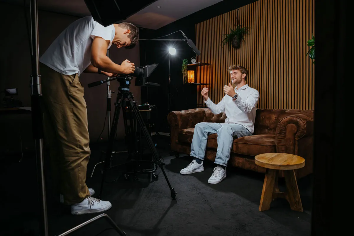 Videographer filming a man sitting on a brown sofa in a studio with professional lighting and microphone equipment.