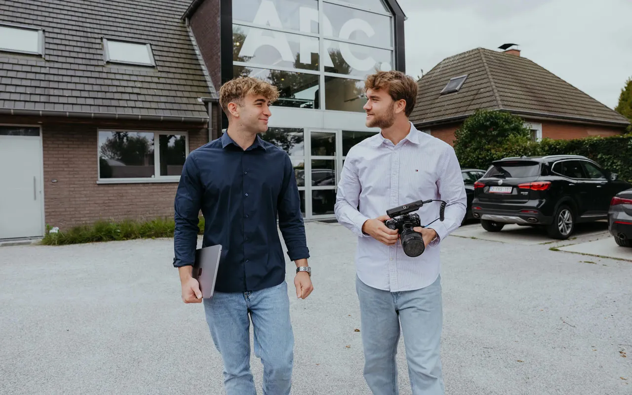 Two young men walking outside a building with ARC signage, one holding a laptop and the other a camera.