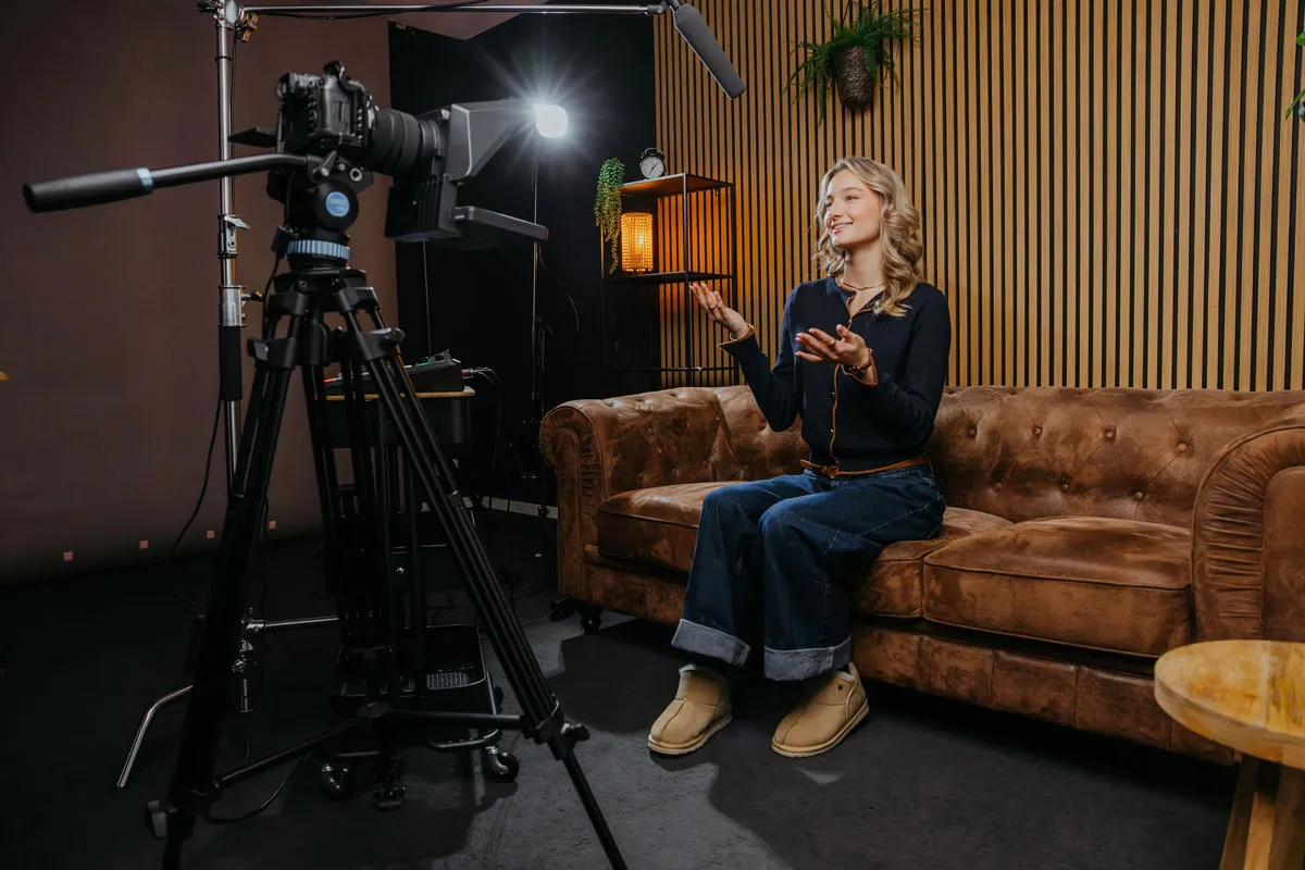 Young woman sitting on a brown leather couch in a recording studio, speaking and gesturing towards a camera on a tripod.