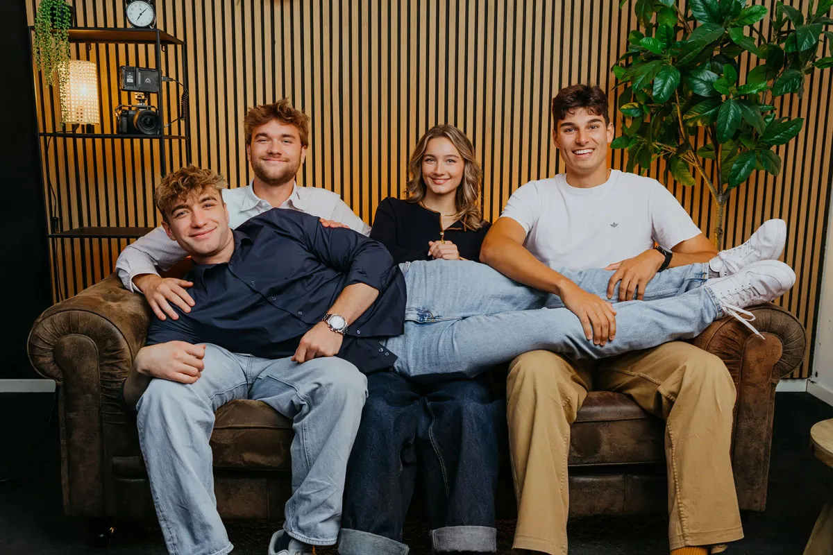 Four young adults smiling on a brown couch, with one lying across the laps of the others in a cozy room with wooden slat wall and green plant.