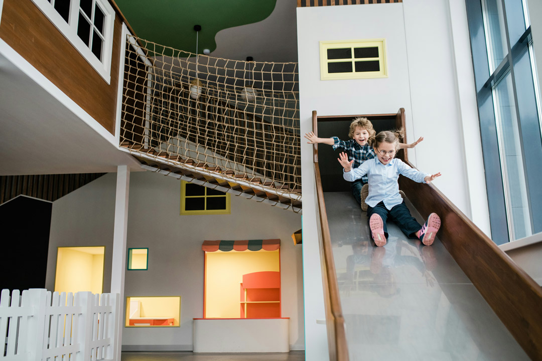 Two children happily sliding down an indoor playground slide with arms outstretched.