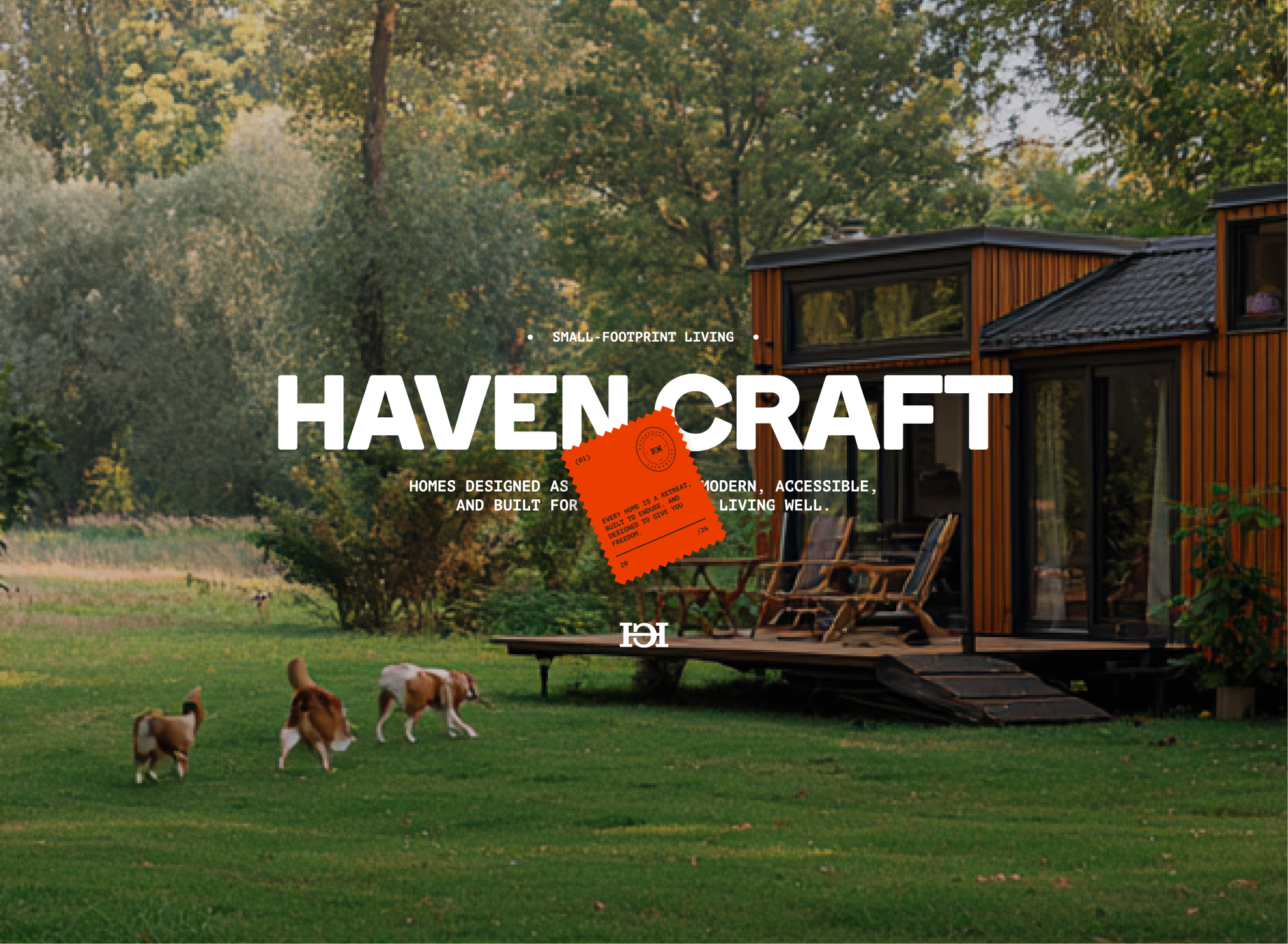 Modern small wooden cabin with a deck and three dogs walking on the green lawn in front surrounded by trees.