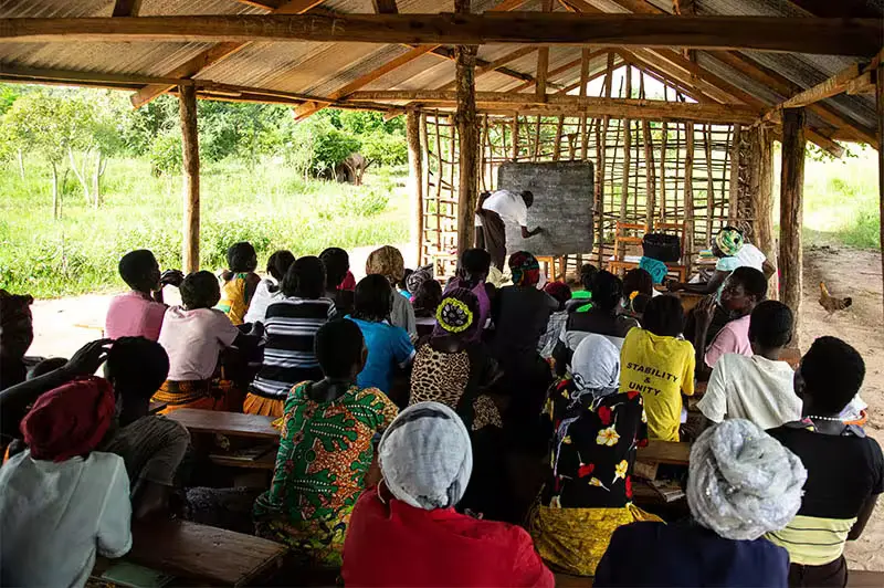 30 local Ugandan women attending an English lesson in a community classroom with a teacher writing on the blackboard