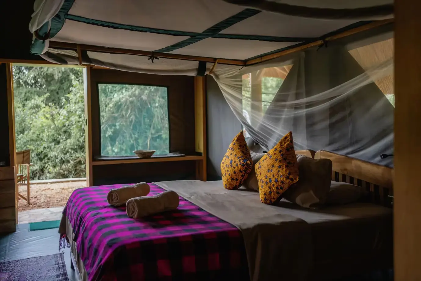 Interior of en-suite Thatched Safari Tent with a double bed, showcasing neatly arranged pillows and rolled towels. The view through the open door reveals a picturesque outdoor setting.