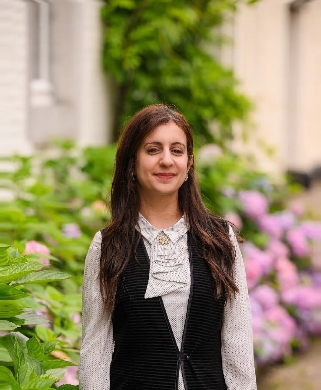 Woman in black vest smiling in garden with pink and green flowers