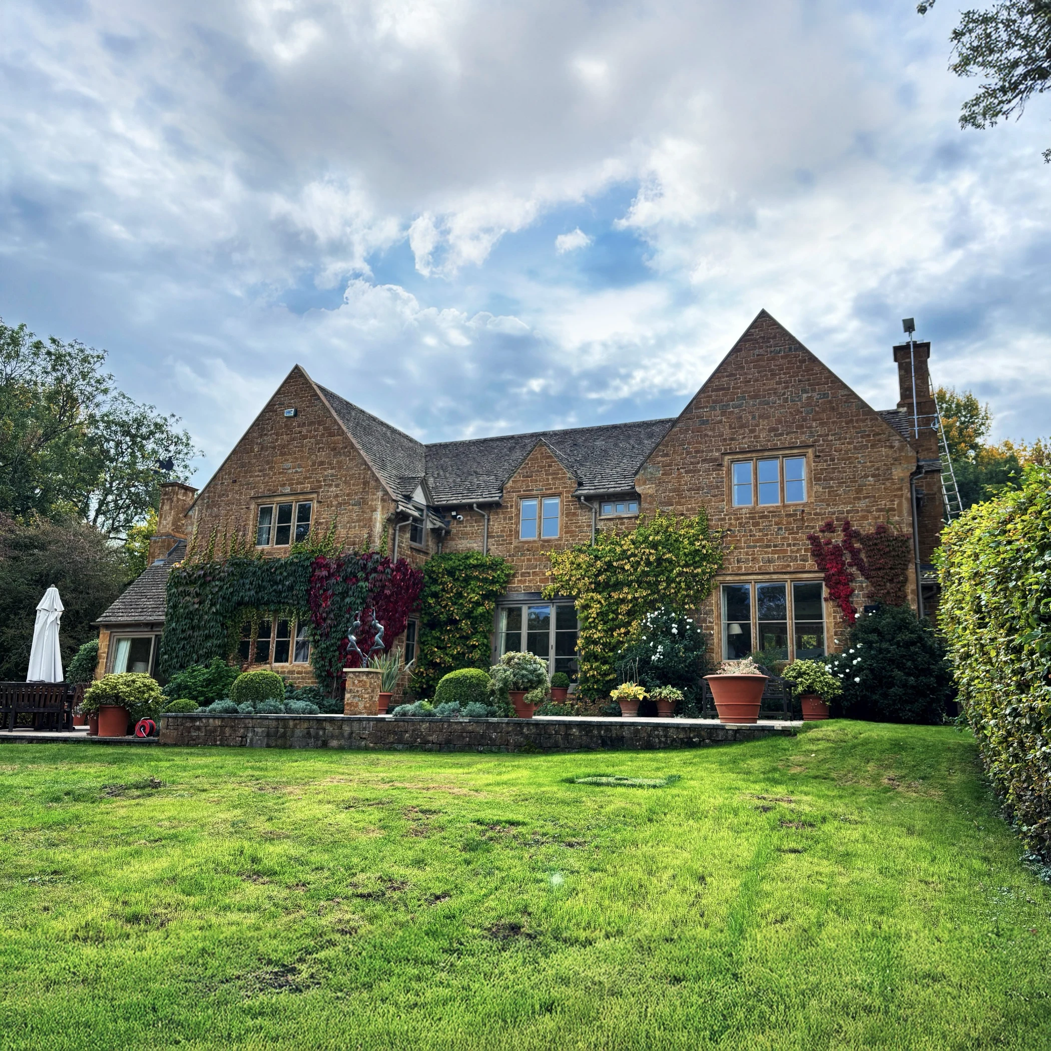 Traditional English country house with warm-toned stone walls, pitched roofs, and large multi-pane windows. The house is surrounded by neatly trimmed gardens, potted plants, and climbing vines with autumn colours, set against a partly cloudy sky.