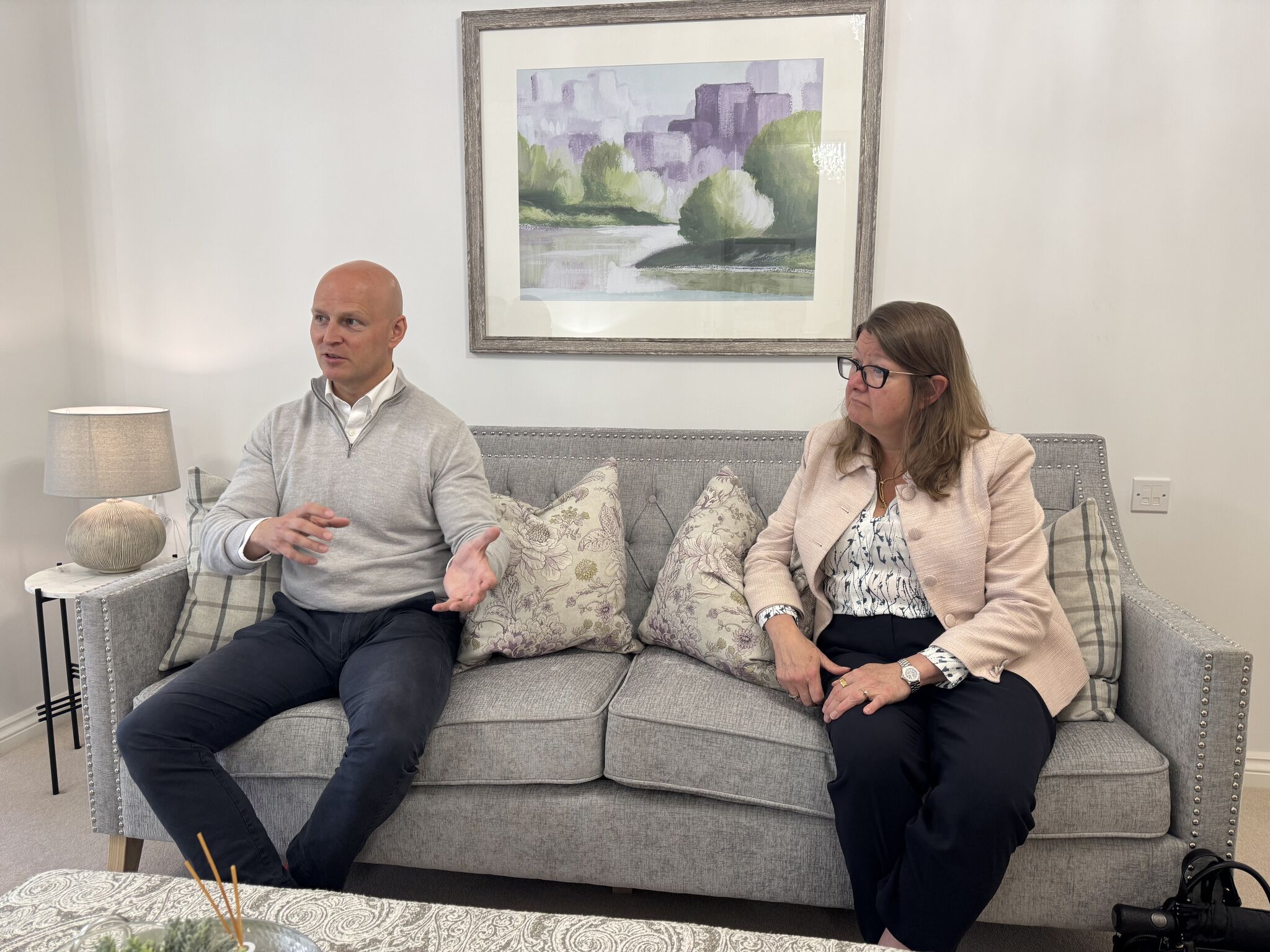 Two people sitting on gray couch with landscape painting in background