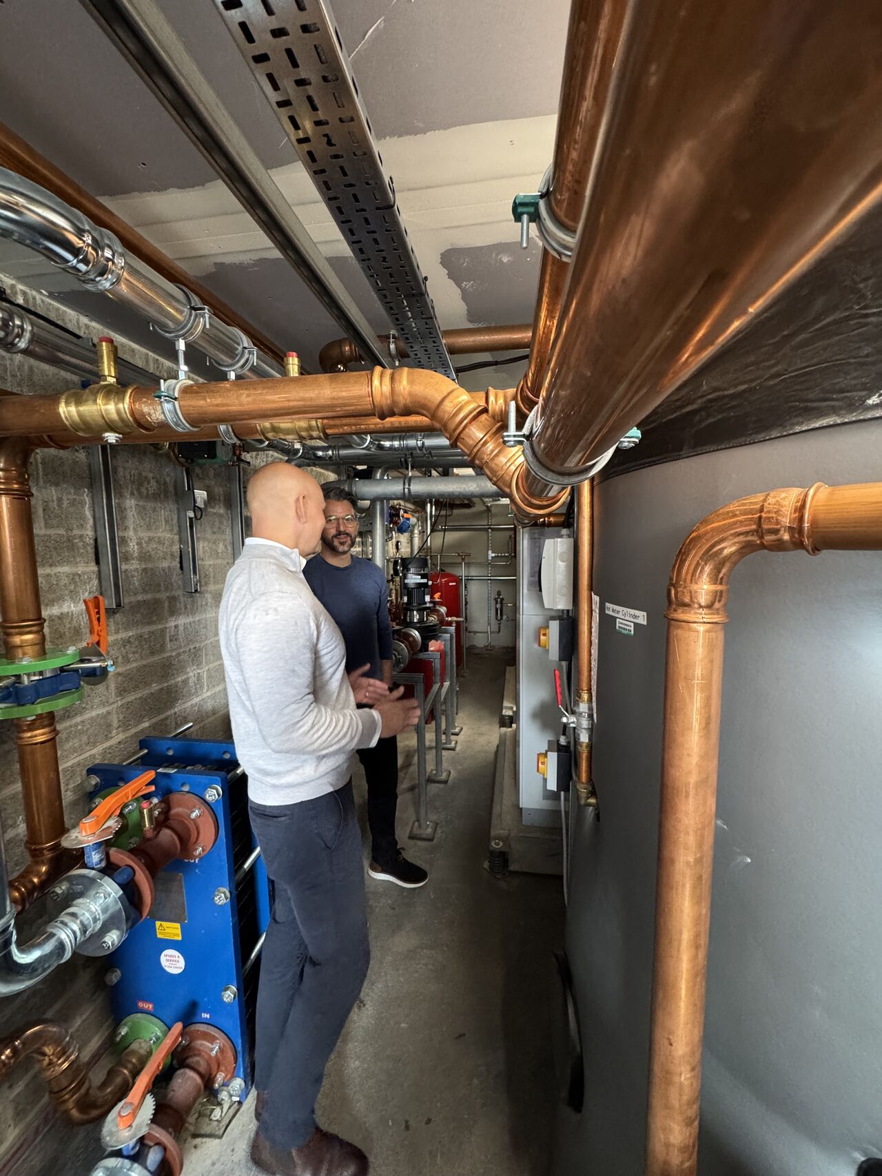 Two technicians inspect copper pipes and mechanical equipment in utility room