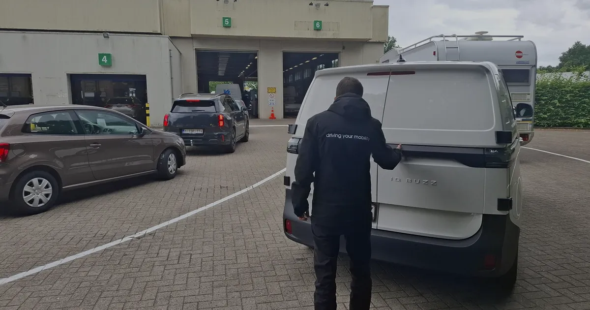 Person in a black jacket opening the rear hatch of a white Volkswagen ID Buzz van in a parking lot with other cars and a building with numbered garage doors.