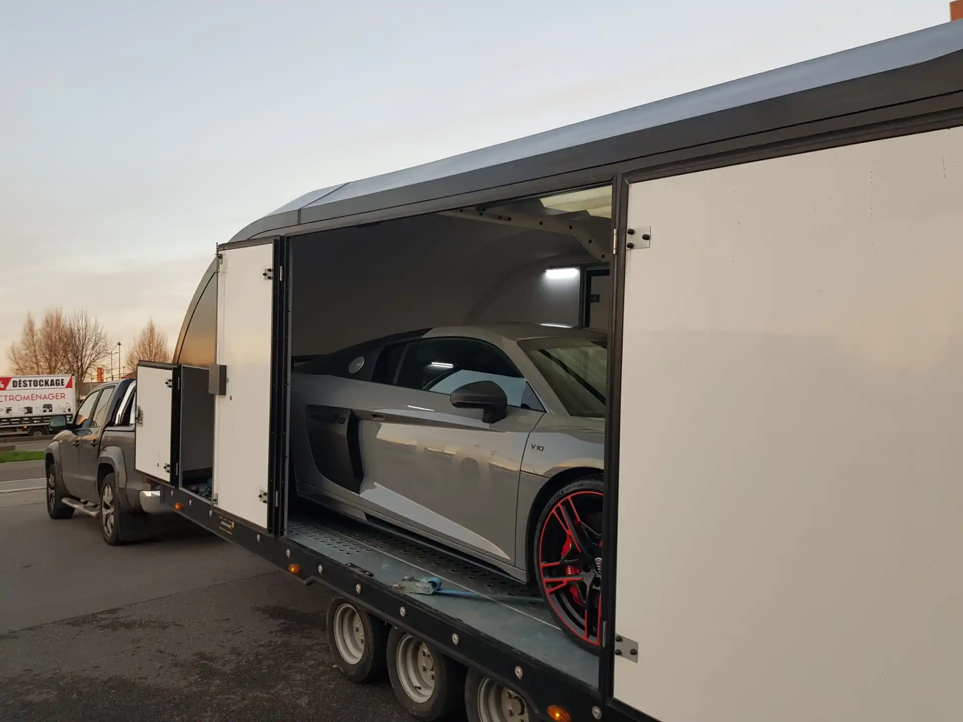 Grey Audi R8 sports car with red-accented wheels inside an open enclosed trailer hitched to a grey pickup truck.
