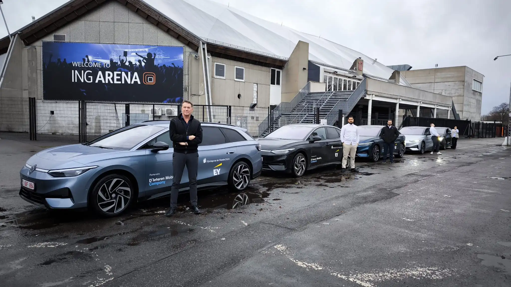 Five people standing next to a line of five parked Volkswagen electric cars outside ING Arena on a cloudy day.