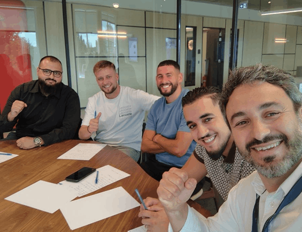 Five men sitting around a wooden table with papers and pens, smiling and giving thumbs up or fist gestures in a modern office setting.