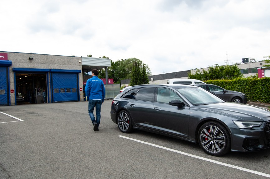 Man in blue jacket walking near a gray station wagon parked outside a garage with blue doors.
