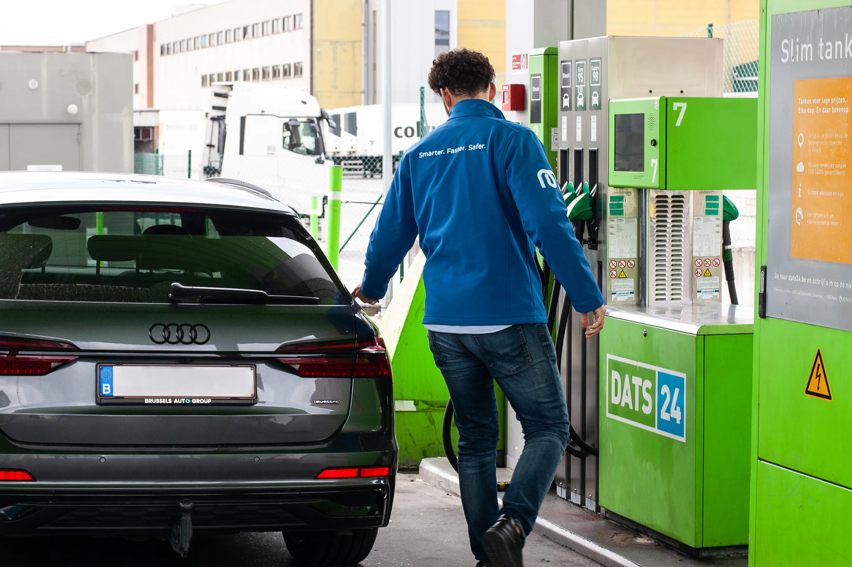 Man in blue jacket fueling a dark gray Audi e-tron at a green DATS 24 gas station pump.
