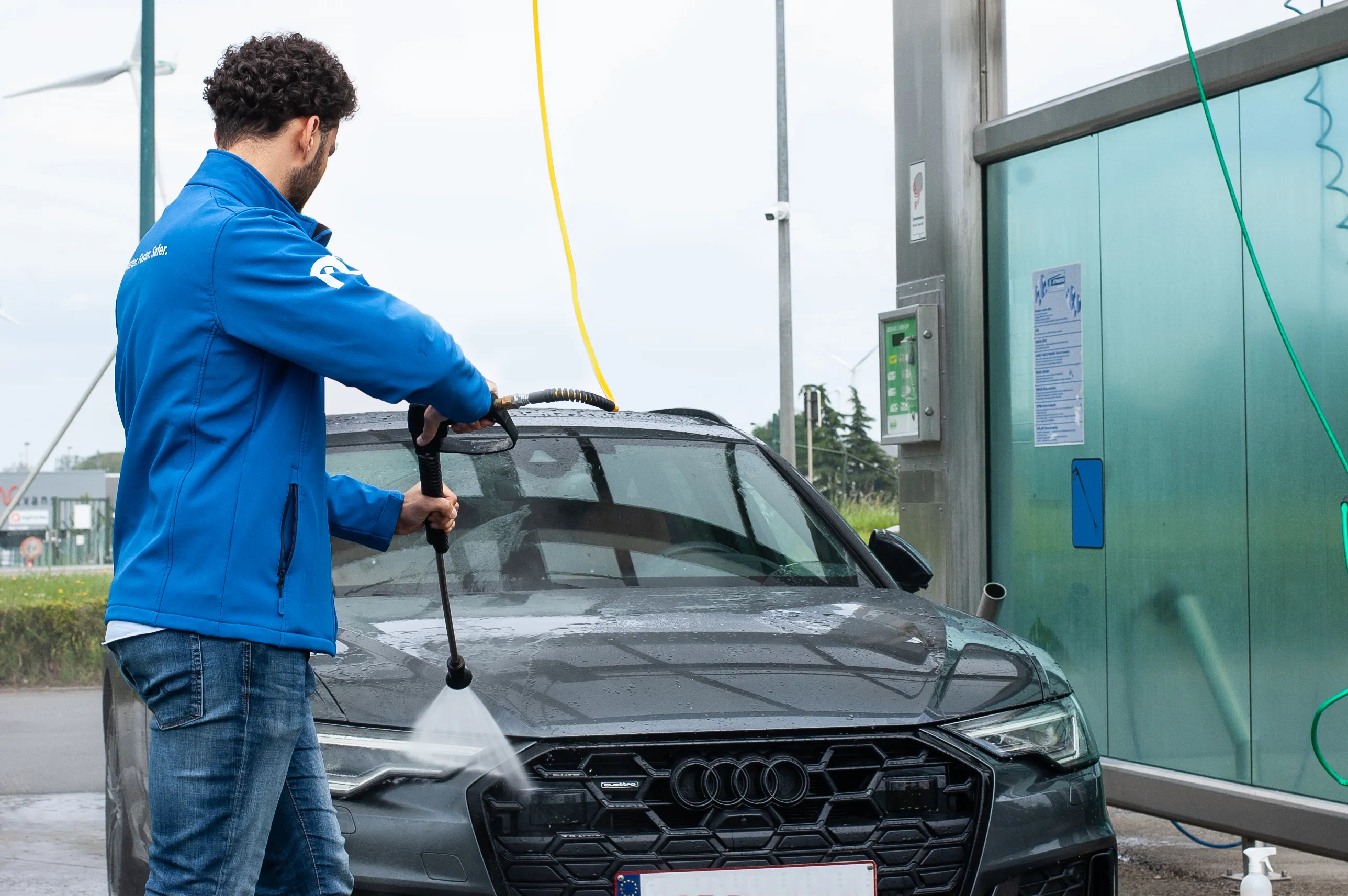 Man in blue jacket cleaning the front of a black Audi car with a pressure washer.
