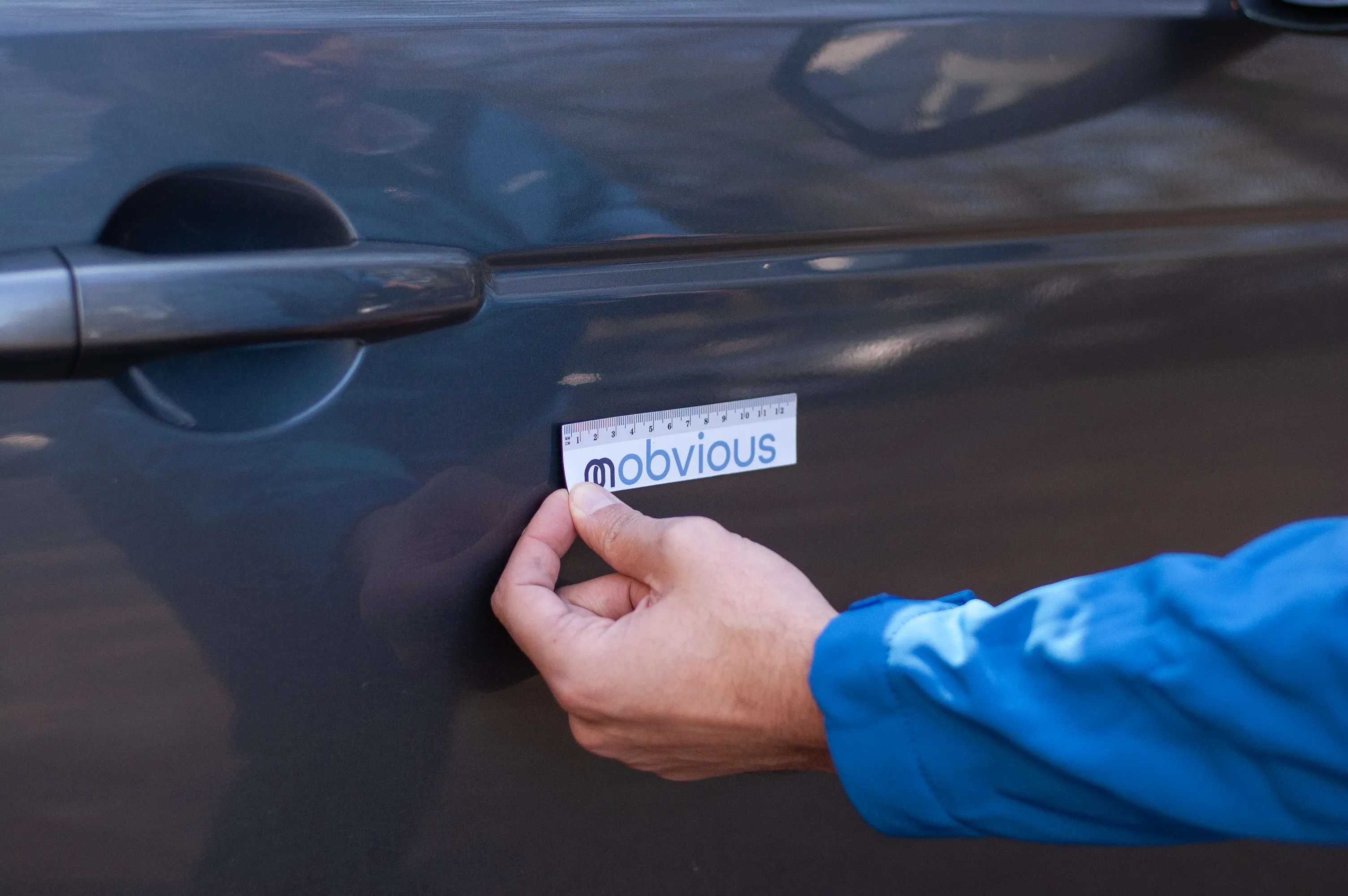 Person holding a blue-and-white ruler labeled 'mobvious' against a dark car door near the handle.