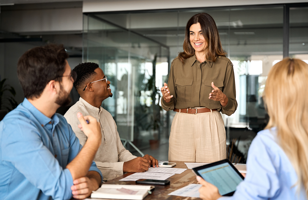 Four diverse colleagues engaged in a lively office meeting, with a woman standing and speaking while others listen and take notes.