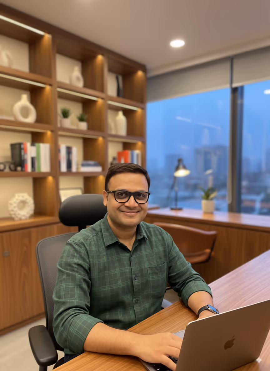 CEO of Truffle Wealth, an Indian man in glasses and a green checked shirt, smiling while working on a laptop in a modern office with bookshelves and a city view in the background